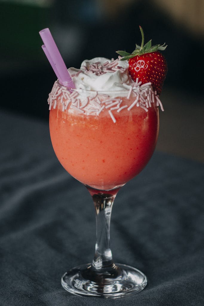 Delicious strawberry smoothie garnished with whipped cream and sprinkles in a wine glass.