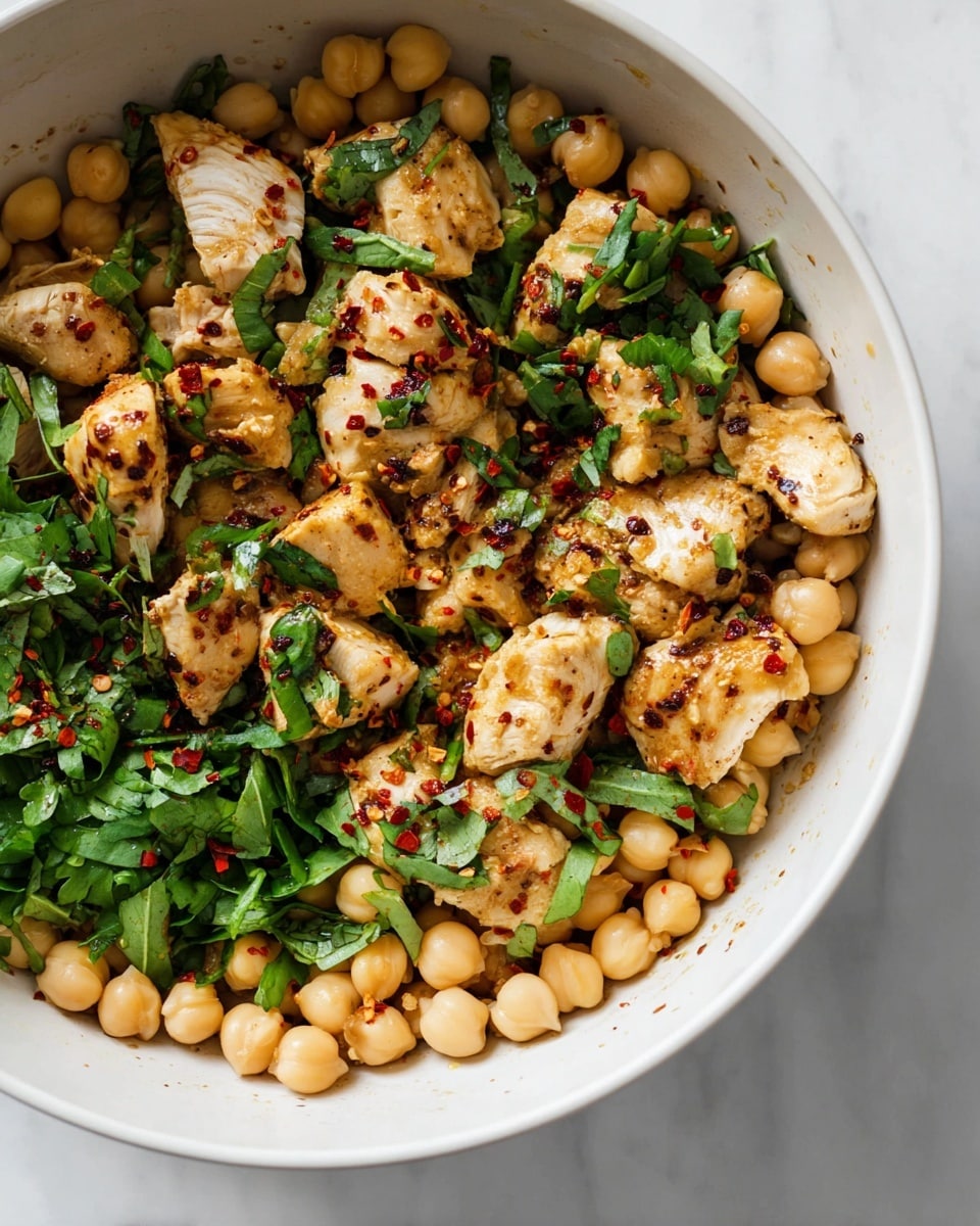 The image shows a white pan with cooked food inside, placed on a white marbled surface. The dish consists of two main layers: the bottom layer is made up of round, light brown chickpeas, scattered evenly across the pan. The second layer is pieces of cooked chicken, light tan and slightly browned, resting on top of the chickpeas. Small bits of green vegetable pieces are mixed throughout, adding a touch of color and texture. The pan has some reddish-brown seasoning sprinkled around the food and the inner sides of the pan. The pan handle is visible at the top right corner. photo taken with an iphone --ar 4:5 --v 7
