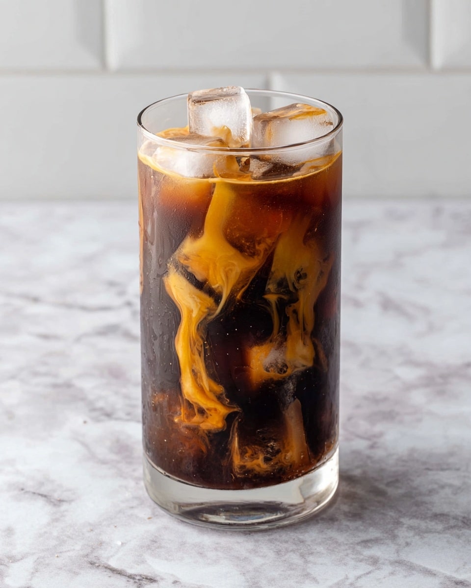 A tall clear glass filled with dark brown cold coffee, with several large ice cubes floating on top. Drips and swirls of golden caramel sauce are visible inside the glass, creating a marbled effect over the coffee. The glass sits on a white marbled surface with a white tiled wall in the background. photo taken with an iphone --ar 4:5 --v 7
