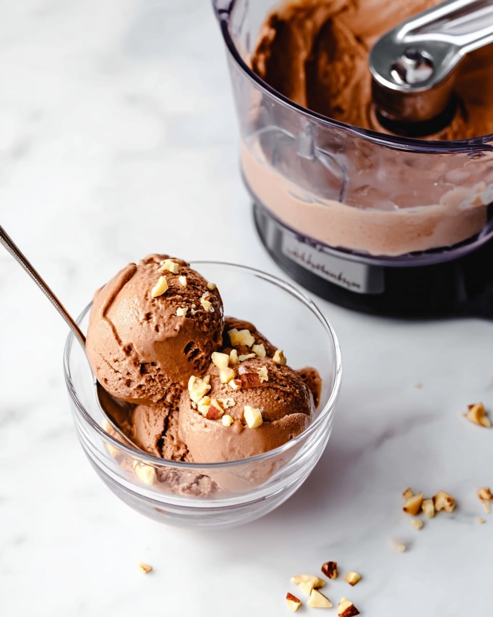 Two rounded scoops of creamy chocolate ice cream with a smooth texture sit in a clear glass bowl, topped with small chopped nuts scattered on and around the ice cream. A silver spoon rests inside the bowl beneath the ice cream. Next to the bowl is a transparent ice cream maker container filled with remaining chocolate ice cream, showing its soft and creamy surface. The scene is set on a white marbled surface with minimal background items to keep focus on the ice cream and bowl. photo taken with an iphone --ar 4:5 --v 7