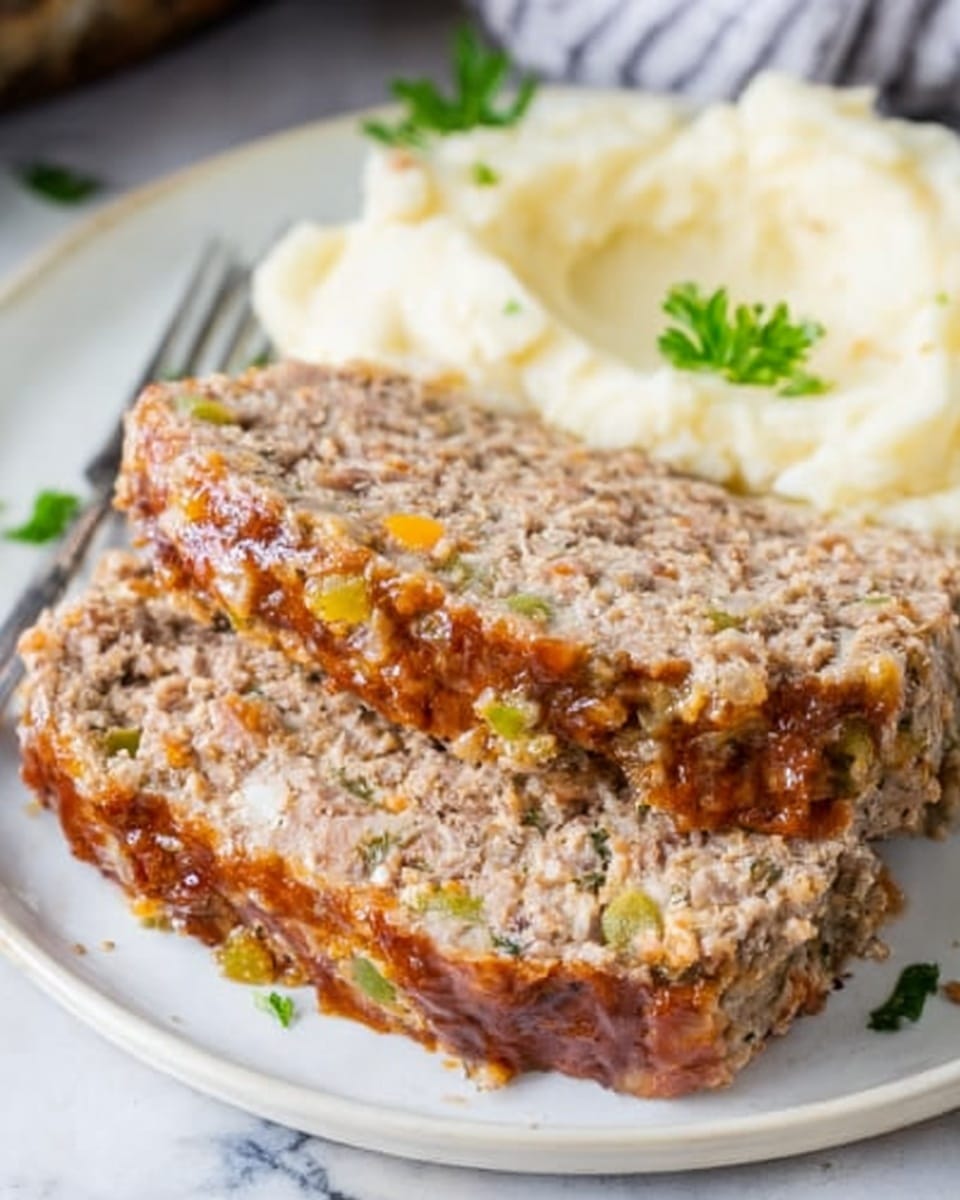 A thick slice of meatloaf with a rough texture and small visible pieces of green and orange vegetables is positioned on the left side of a white plate, showing a slightly glossy top edge with a reddish glaze. To the right, there is a portion of creamy mashed potatoes sprinkled with a few green herbs. A silver fork rests on the plate near the mashed potatoes, holding a small piece of meatloaf. The background includes blurred green herbs on a white marbled surface, and a cutting board with more meatloaf is visible out of focus. Photo taken with an iphone --ar 4:5 --v 7