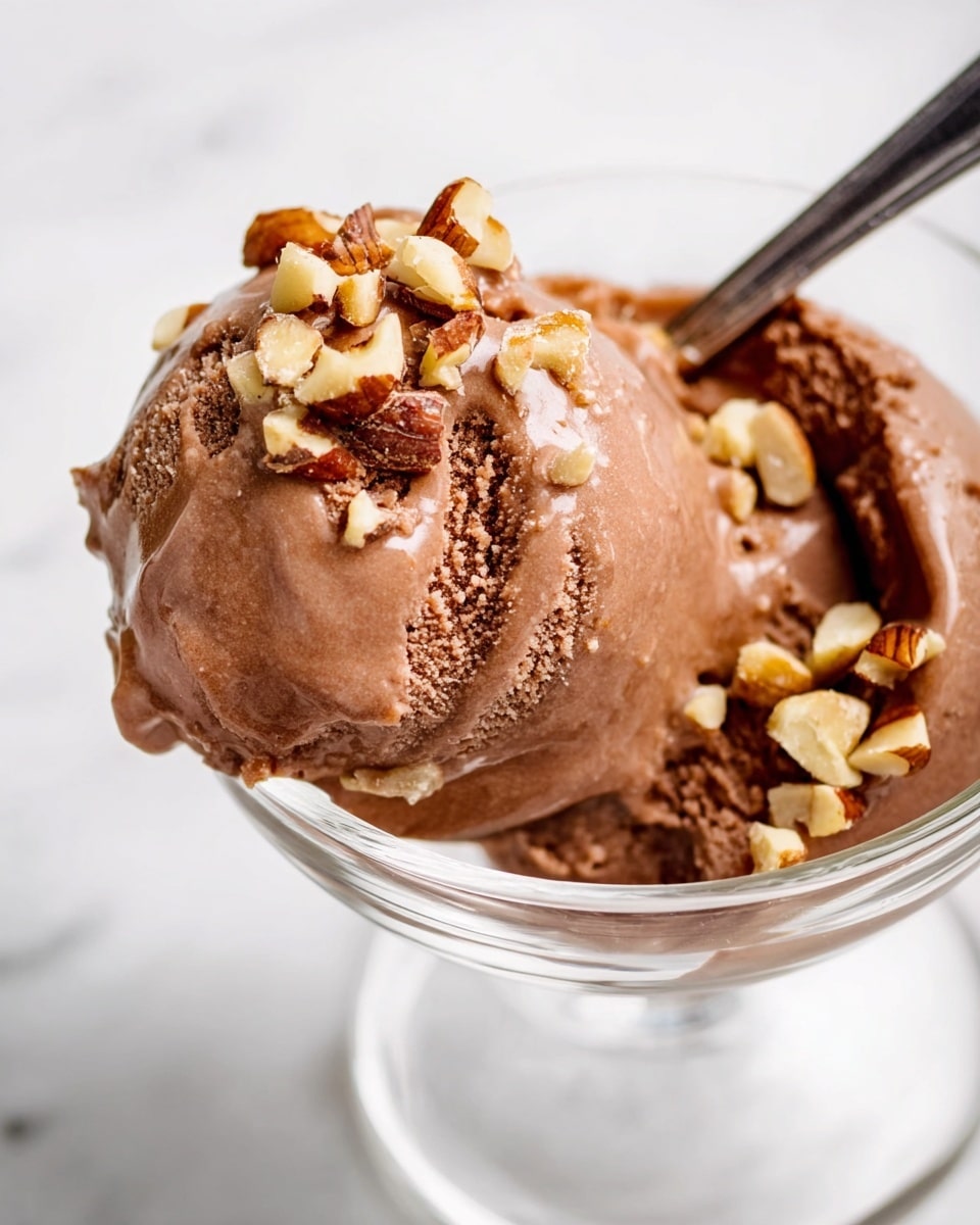 A close-up view shows one scoop of smooth, creamy chocolate ice cream with a rich brown color sitting in a clear glass bowl. The ice cream is topped with small pieces of chopped nuts that add a crunchy texture with shades of light beige and brown. Some ice cream and nuts rest on the silver spoon placed inside the bowl, and the background features a white marbled texture. photo taken with an iphone --ar 4:5 --v 7