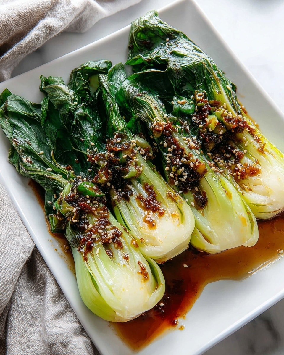 The image shows four cooked baby bok choy pieces arranged neatly in a row on a white rectangular plate. Each piece has a vibrant green leafy base that looks soft and wilted, transitioning to a glossy, light green to pale yellow stalk that appears tender and slightly charred. The bok choy is topped with a shiny, dark brown sauce containing tiny bits of red chili and garlic, sprinkled with toasted sesame seeds for texture. The plate rests on a white marbled surface, with a soft cloth in the background. photo taken with an iphone --ar 4:5 --v 7