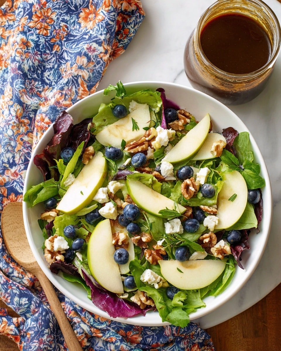 A fresh salad is shown in a white bowl on a white marbled surface, layered with mixed green and purple leafy lettuce as the base. Thin slices of light green apple are scattered on top, along with deep blue blueberries and small chunks of white cheese. Pieces of brown walnut are spread throughout, and the salad is finished with green herb leaves sprinkled over everything. To the side, there is a jar filled with dark brown dressing, and part of a cloth with blue and orange floral patterns is visible. photo taken with an iphone --ar 4:5 --v 7