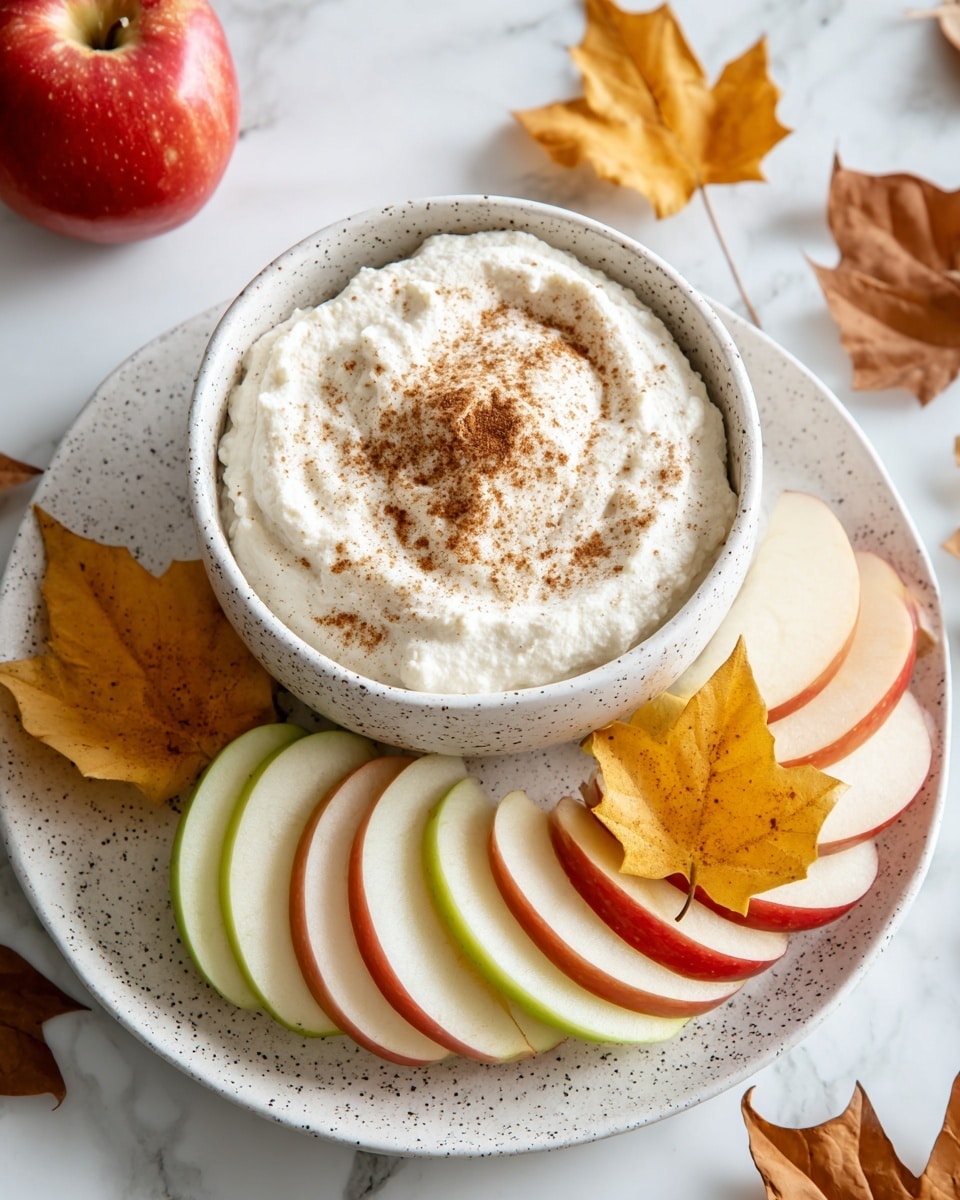 A white speckled bowl filled with creamy white dip sprinkled with brown spice on top, placed in the center of a white speckled plate. Around the bowl, there are thin slices of red and green apples, neatly arranged in a curved line. The plate also has a few yellow and orange autumn leaves scattered near the apple slices. The setting is on a white marbled surface with a whole red apple and some more autumn leaves visible in the background. Photo taken with an iphone --ar 4:5 --v 7