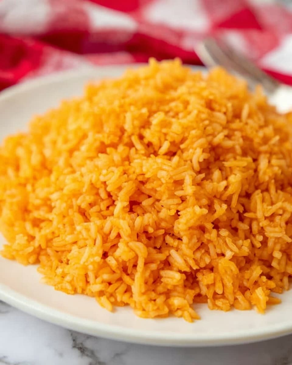 A close-up of a pile of fluffy orange rice served on a white plate, each grain distinct and coated with a soft, slightly shiny sauce giving it a moist texture, the rice color varies slightly from deeper to lighter orange shades, the plate sits on a white marbled surface with a blurred red and white checkered cloth and a fork in the background, photo taken with an iphone --ar 4:5 --v 7
