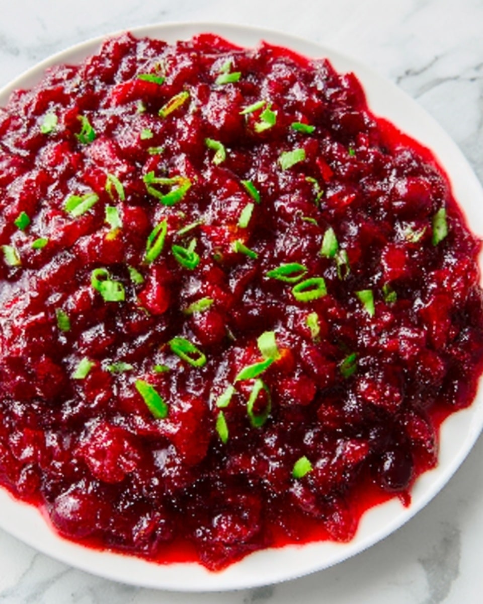 The image shows a close-up of a dish in a white plate filled with a deep red chunky mixture that looks like cooked cranberries or a berry sauce. The top layer is glossy with visible pieces of berries and is sprinkled with small green chopped herbs evenly scattered on top, adding a fresh contrast. The texture looks thick and slightly sticky, with some crushed berry bits visible throughout. The background surface is a white marbled texture which highlights the rich color of the dish. Photo taken with an iphone --ar 4:5 --v 7