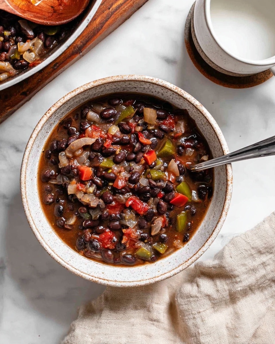 A white speckled bowl filled with a thick stew made of dark black beans, chopped green peppers, large pieces of translucent cooked onions, and chunks of red tomatoes mixed in a glossy, dark broth. A silver spoon is partially submerged on the right side inside the bowl. The bowl sits on a white marbled surface, next to a beige cloth on the bottom right and a white speckled dish with a white cup filled with a white liquid on the top right. The edge of a white pan filled with the same stew and a wooden spoon holding cooked onions and beans is visible on the top left. Photo taken with an iphone --ar 4:5 --v 7