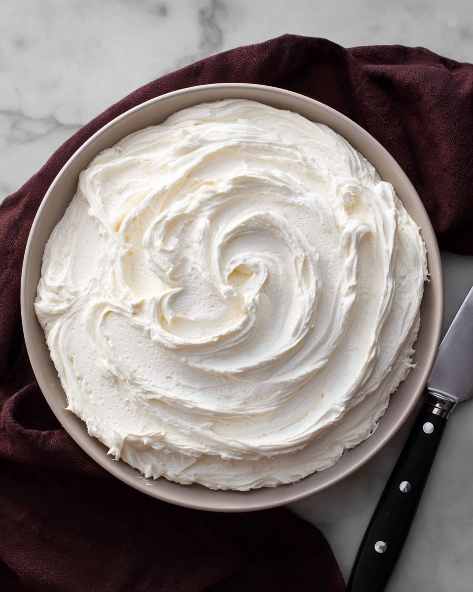 A close-up top view of a thick, creamy white frosting spread in generous, smooth swirls inside a round white bowl, filling it nearly to the edge with soft peaks and a whipped texture. The bowl sits on a rich white marbled surface with a dark burgundy cloth partially tucked under it. To the right, a black-handled spatula with a shiny metal blade rests on the surface. The overall look is clean and inviting with soft light highlighting the texture of the frosting. photo taken with an iphone --ar 4:5 --v 7