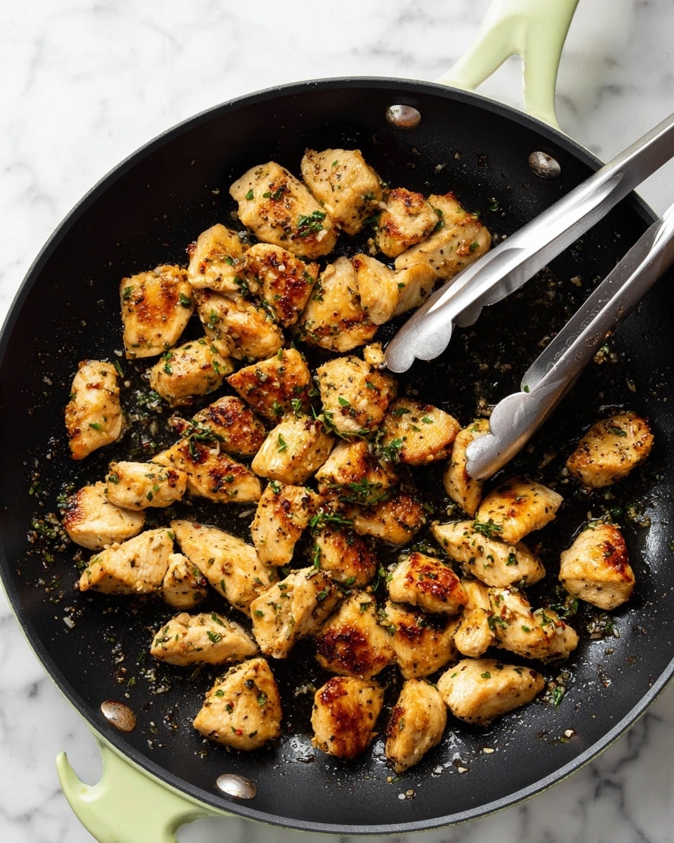 The image shows a pan filled with small pieces of golden-brown cooked chicken. Each piece has a crispy, slightly charred outside with a juicy, tender texture inside. The chicken is scattered unevenly across the black pan and is sprinkled with small bits of green herbs. There are shiny spots of oil and tiny garlic pieces visible on the pan surface. A pair of silver and black tongs is holding some chicken near the top right of the pan. The pan is sitting on a white marbled surface. photo taken with an iphone --ar 4:5 --v 7