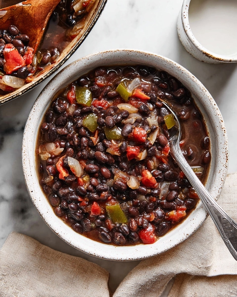The image shows a large stainless steel pan filled with a cooked black bean stew. The stew contains layers of dark black beans mixed with diced translucent white onions and soft green bell peppers, scattered throughout. There are pieces of cooked red tomatoes adding spots of bright color. The stew looks thick and hearty with some liquid sauce surrounding the beans and vegetables. A wooden spoon rests inside the pan, partially covered with sauce and vegetables. To the side, a white bowl with black speckles sits on a white marbled surface next to a light beige cloth. Photo taken with an iphone --ar 4:5 --v 7