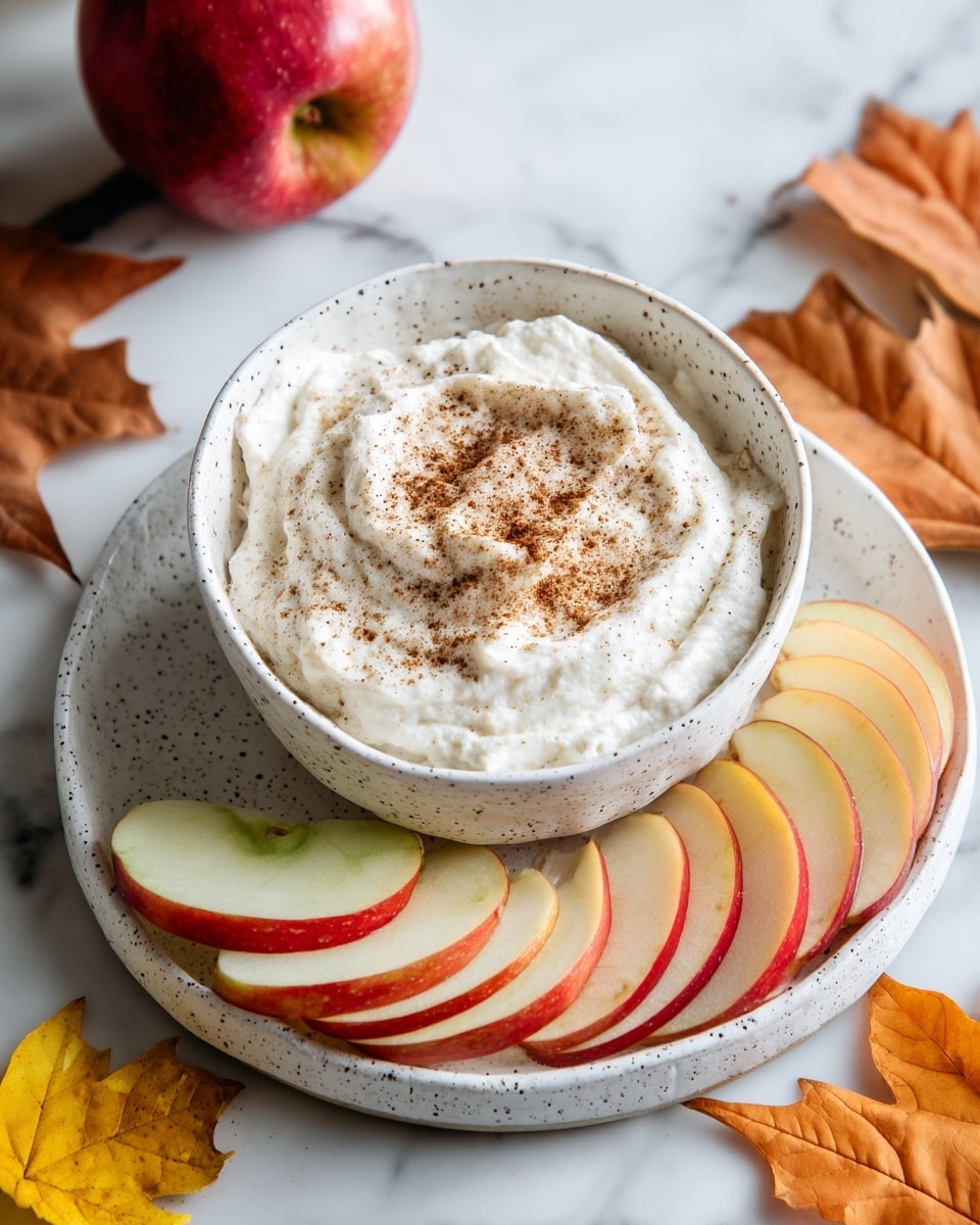 A white speckled bowl filled with a thick, creamy white dip topped with a light dusting of brown cinnamon powder sits in the center of a white speckled plate. Around the bowl on the plate are alternating slices of red and green apple, arranged in a neat overlapping row. Three yellow and brown dried maple leaves are placed decoratively on the plate near the apple slices. The plate rests on a white marbled surface with a whole red apple and additional dried leaves scattered around. photo taken with an iphone --ar 4:5 --v 7