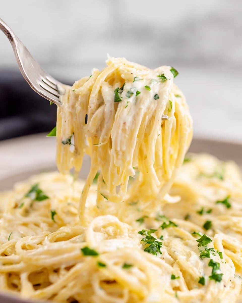 A close-up image of creamy spaghetti lifted by a fork, showing long, smooth noodles covered in a thick, white sauce. Small green herbs are scattered on and between the noodles, adding a pop of color. The background is softly blurred, focusing all attention on the glossy texture of the pasta and sauce. The dish sits on a surface with a white marbled texture. Photo taken with an iphone --ar 4:5 --v 7