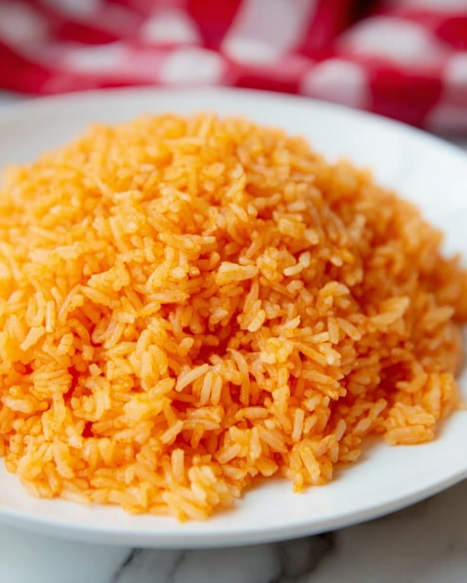 A close-up view of a mound of bright orange rice served on a white plate, showing the fluffy, separate grains evenly colored with a soft texture. The rice has a glossy look with slight variations in the orange shade, indicating it is well cooked and seasoned. The plate sits on a white marbled surface with a blurred red and white checkered cloth in the background, adding a cozy feel. Photo taken with an iphone --ar 4:5 --v 7