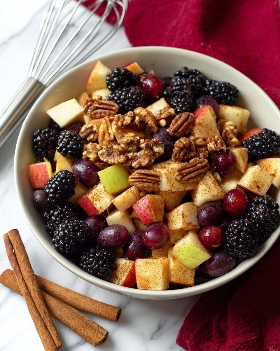 The image shows a white bowl filled with a fresh fruit salad made of three main layers: shiny red grapes scattered throughout, blackberries with a bumpy texture filling the gaps, and diced apple chunks with red skin and yellow flesh coated lightly in cinnamon. Mixed in are small pieces of walnuts and pecans adding a rough texture. The bowl is placed on a white marbled surface, with two cinnamon sticks beside it. A metal whisk and a red and beige patterned cloth are visible in the background. photo taken with an iphone --ar 4:5 --v 7