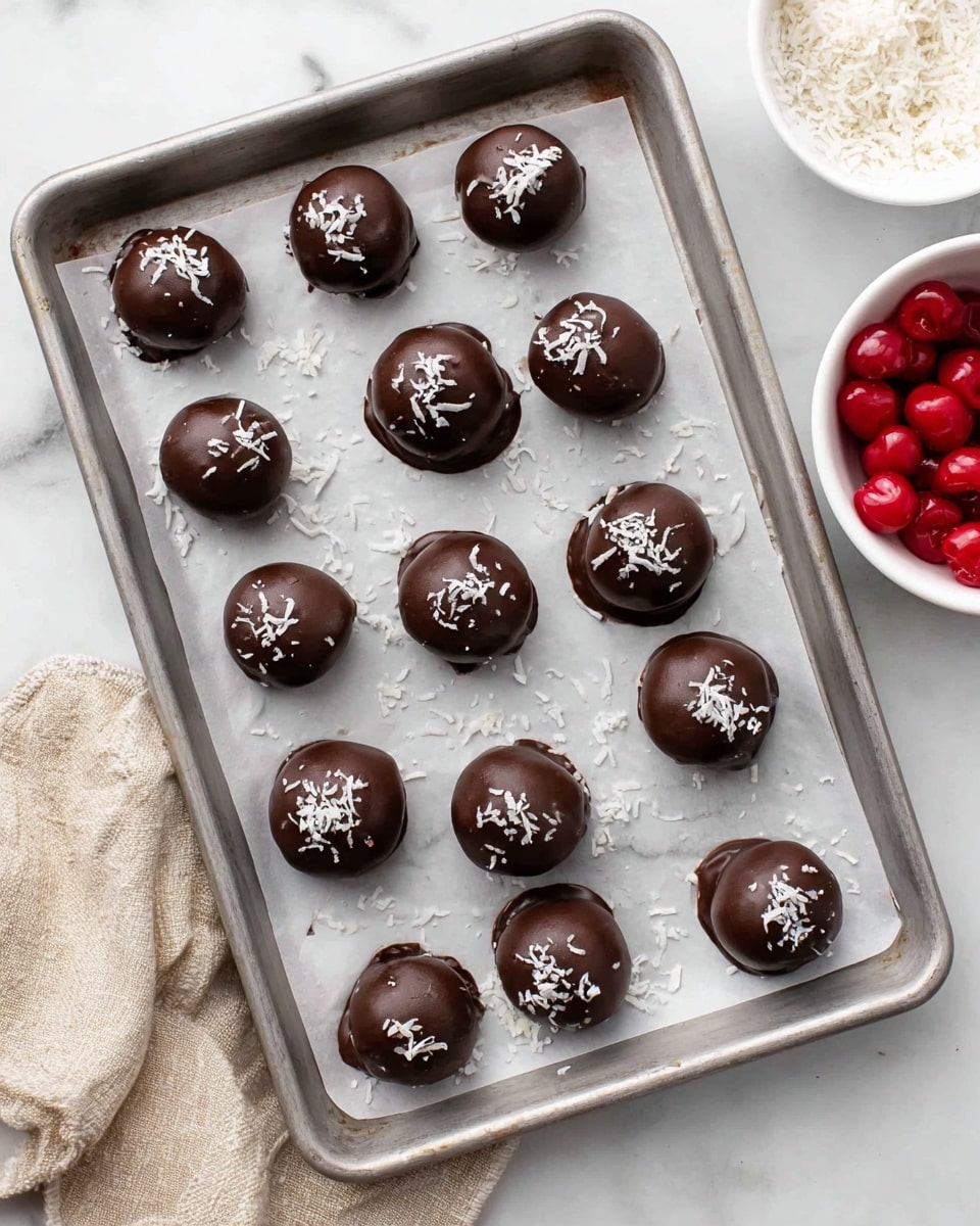A metal baking tray lined with white parchment paper holds fifteen round chocolate-covered balls. Each ball is smooth and shiny, colored dark brown from the chocolate shell, with some topped with small white coconut flakes. The balls are arranged loosely in rows, with a few loose coconut flakes scattered around them on the parchment. On the right side, there is a small white bowl filled with bright red cherries, and part of another white bowl is visible at the top edge. The whole scene is set on a white marbled surface with a beige cloth partially visible in the lower left corner. Photo taken with an iphone --ar 4:5 --v 7
