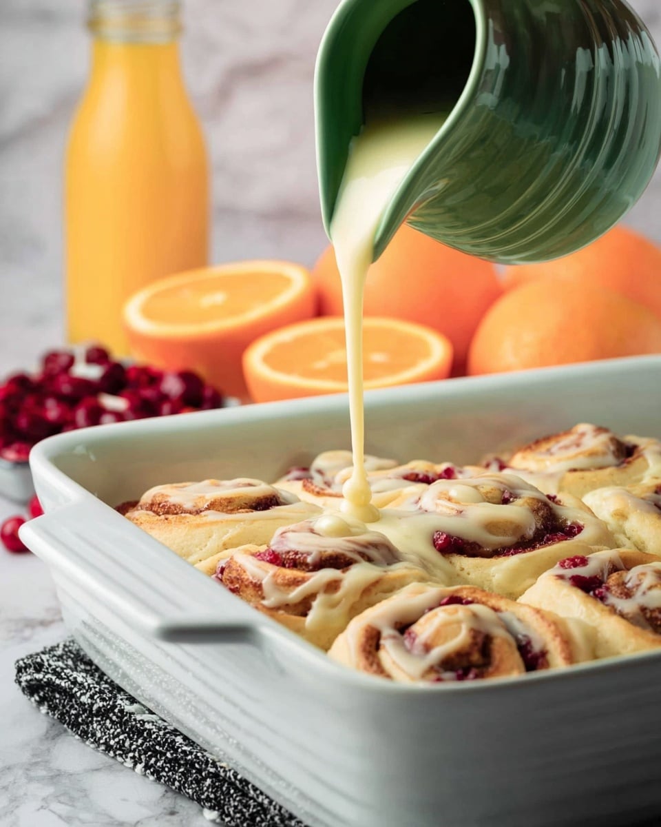 A close-up of a soft, partially eaten cinnamon roll layered with creamy white icing on top, revealing a gooey, bright red cranberry filling inside. The roll shows a light beige dough with visible swirls of red fruit sauce and plump cranberries around and within the layers. It sits on an elegant white plate with a black fork resting beside it, holding a piece of the roll with cranberry. In the background, there are slices of bright orange and a white bowl filled with fresh cranberries, all placed on a white marbled surface. photo taken with an iphone --ar 4:5 --v 7