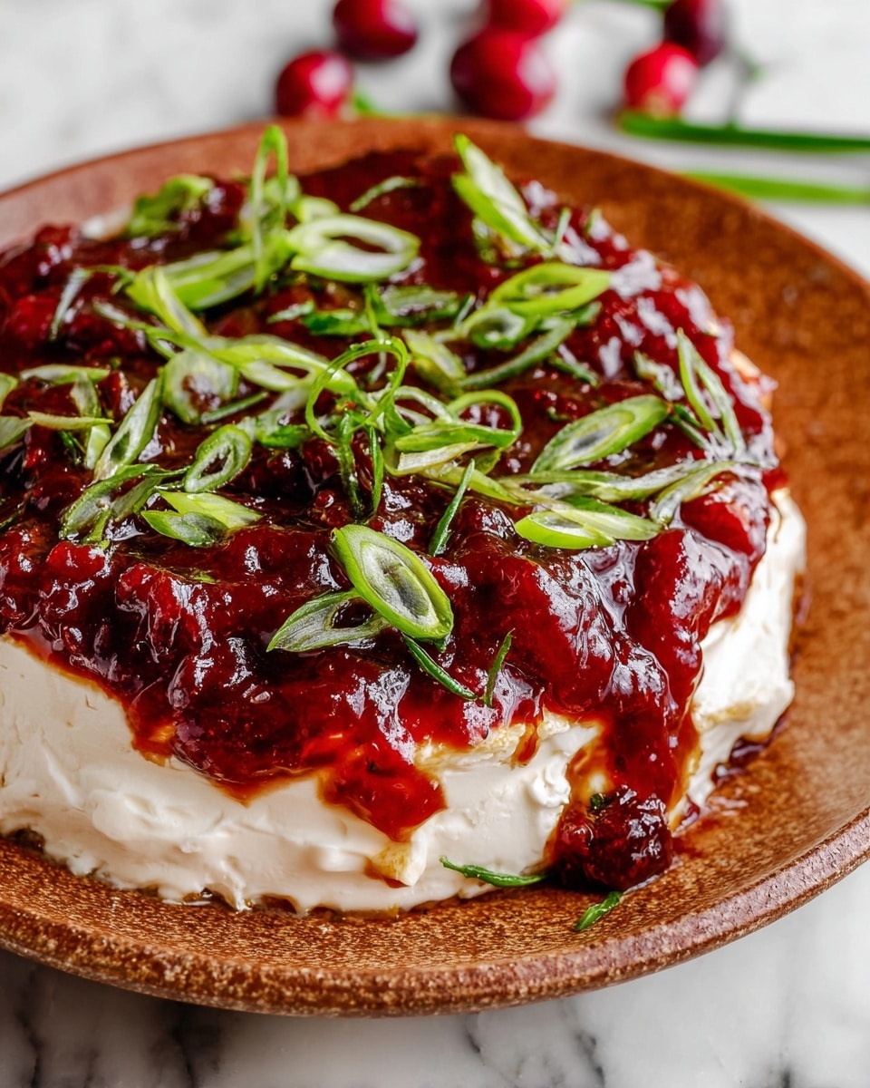 A round beige bowl holds a two-layer dip on a wooden cutting board. The bottom layer is a creamy white spread with a soft texture, topped with a thick, glossy, deep red cranberry sauce. Fresh green chopped scallions are scattered across the red layer, adding a fresh contrast. To the right side of the bowl, there are many square beige crackers arranged loosely. The wooden board has a worn texture, placed on a white marbled surface. photo taken with an iphone --ar 4:5 --v 7