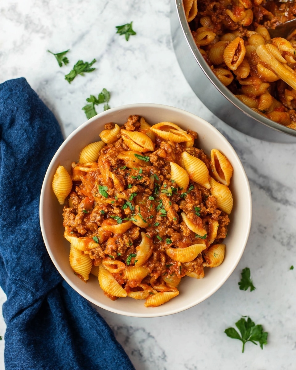 The image shows a close-up of a bowl filled with shell-shaped pasta mixed with browned ground meat and bits of herbs. The pasta is a warm yellow-orange color, covered lightly with a sauce that gives it a glossy look. The ground meat pieces are brown and crumbly, evenly mixed between the pasta shells. Small green herb pieces are scattered on top, adding a touch of color. The food is served in a white bowl with a soft scalloped edge, placed on a white marbled surface with a blurred dark blue cloth in the background. photo taken with an iphone --ar 4:5 --v 7