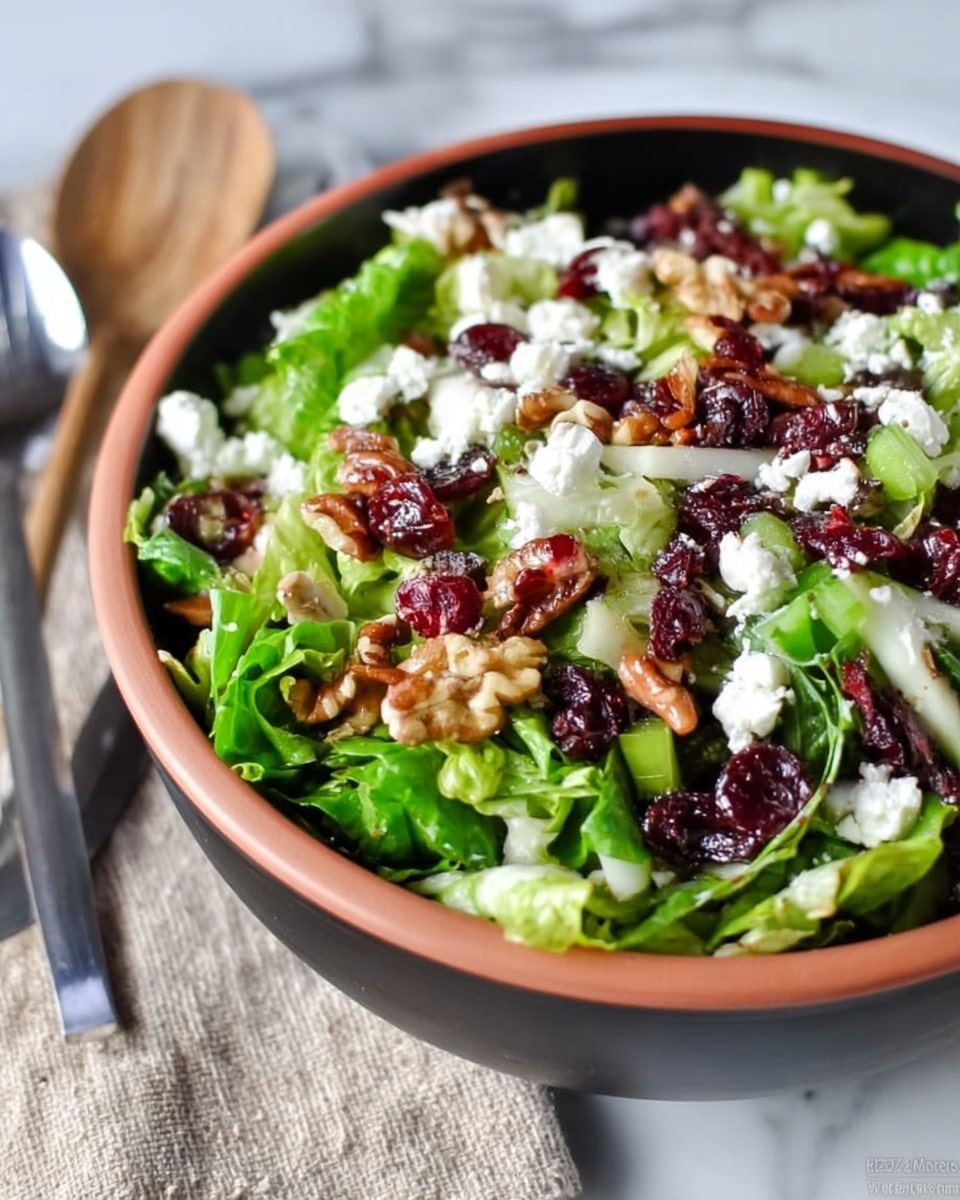 The image shows a salad in a deep white bowl filled with several layers. The bottom layer is bright green leafy lettuce with a fresh and crinkled texture. On top, there are scattered bits of white soft cheese, small dark red dried cranberries, crunchy brown walnuts, and chopped celery pieces that add a fresh light green color. The bowl sits on a white marbled surface, and a spoon and fork are visible beside it. Photo taken with an iphone --ar 4:5 --v 7