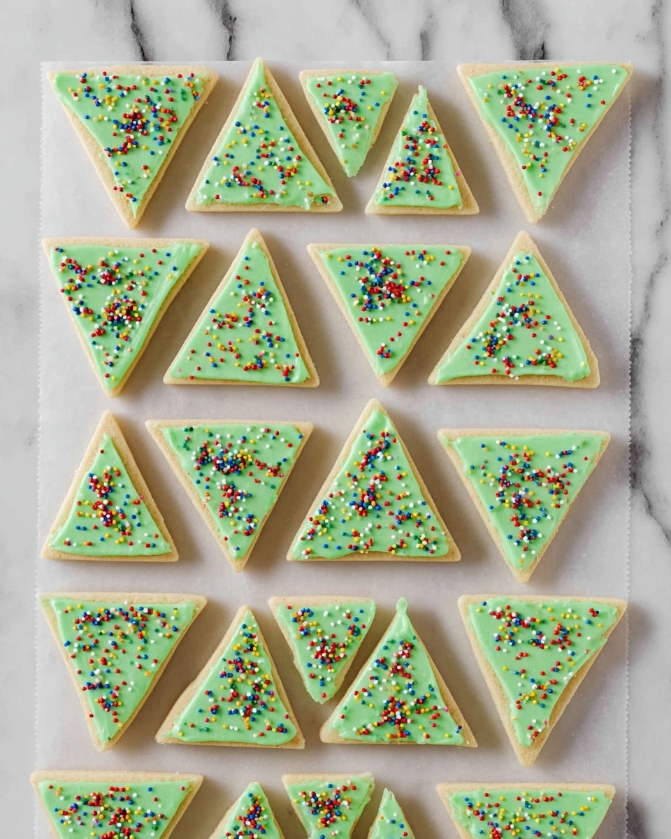The image shows twenty triangular cookies neatly arranged in four rows on a white marbled surface covered with white parchment paper. Each cookie has a pale green frosting layer on top, which is smooth but slightly textured, and is decorated with small, colorful round sprinkles in red, yellow, blue, and pink spread evenly across the surface. The cookies have a light brown base layer visible beneath the frosting, with the frosting layer making up about half the thickness of each cookie. photo taken with an iphone --ar 4:5 --v 7