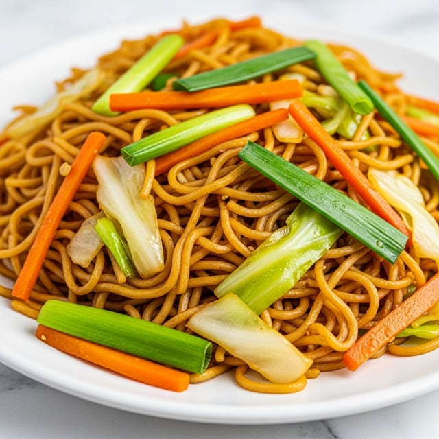 A close-up of a white plate piled high with stir-fried noodles, showing thin, golden-brown noodles mixed with bright orange carrot strips, light green cabbage pieces, and vibrant green scallions. The noodles have a shiny, slightly oily texture, while the vegetables add varied colors and crunchiness. The dish looks warm and freshly cooked with a mix of soft and crispy elements. The plate is set on a white marbled surface. photo taken with an iphone --ar 4:5 --v 7