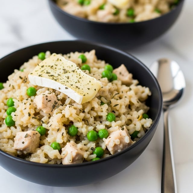 A close-up of a white bowl filled with creamy risotto featuring visible green peas and shredded pieces of light brown chicken, topped with a sprinkle of herbs. The risotto shows a soft, slightly textured surface with small grains and chunks, sitting on a white marbled surface with a silver spoon resting beside the bowl. The background is softly blurred, giving focus to the bowl in the front. photo taken with an iphone --ar 4:5 --v 7