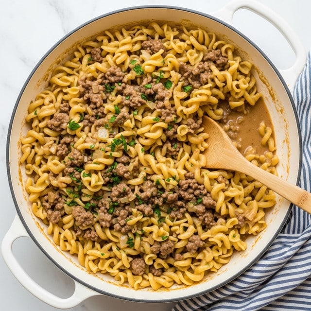 A large white skillet filled with cooked egg noodles mixed with browned ground beef and small chunks of cooked onions, all coated in a light brown, creamy sauce. The noodles are curly and golden-yellow, with small flecks of chopped green herbs sprinkled evenly on top. A wooden spoon rests in the pan, partially submerged in the mixture, giving a close view of the creamy texture and hearty contents. The skillet sits on a white marbled surface next to a blue and white striped cloth. photo taken with an iphone --ar 4:5 --v 7
