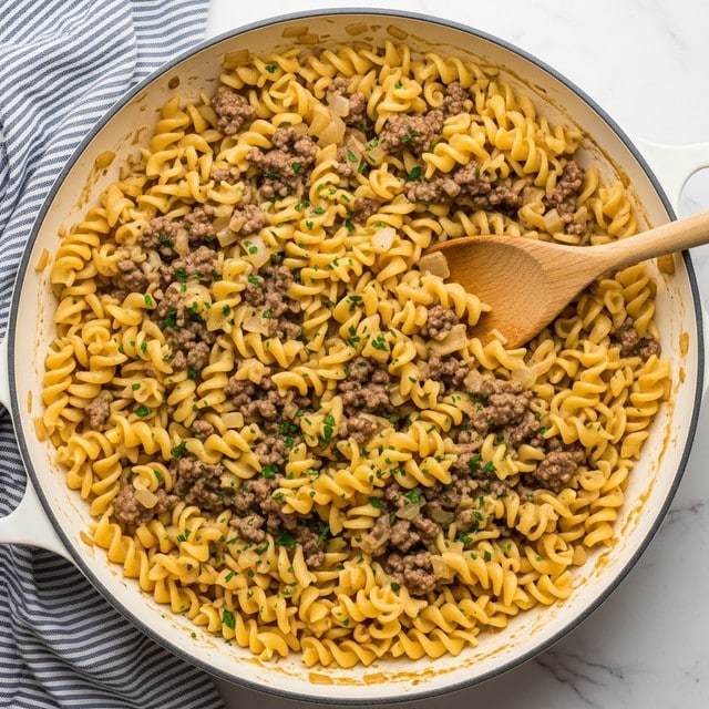 A white deep pan filled with three layers of egg noodles mixed with browned ground beef and chopped onions in a thick light brown sauce, sprinkled with small bits of chopped green parsley on top. The noodles have a soft, twisted texture and a pale yellow color, while the beef pieces are crumbly and dark brown. A wooden spoon is dipped into the pan on the right side, resting against the noodles and sauce. The pan handle and a blue and white striped cloth napkin are on the white marbled surface beneath. photo taken with an iphone --ar 4:5 --v 7
