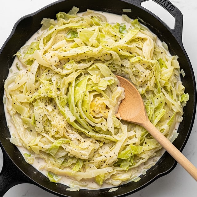 A close-up view of a black cast iron skillet filled with creamy cooked cabbage. The dish has soft, light green and pale yellow cabbage layers mixed with a thick, white cream sauce that coats the cabbage evenly, with small flecks of black pepper scattered on top. A light brown wooden spoon is resting inside the skillet, partially submerged in the creamy mixture. The skillet is placed on a white marbled surface. photo taken with an iphone --ar 4:5 --v 7