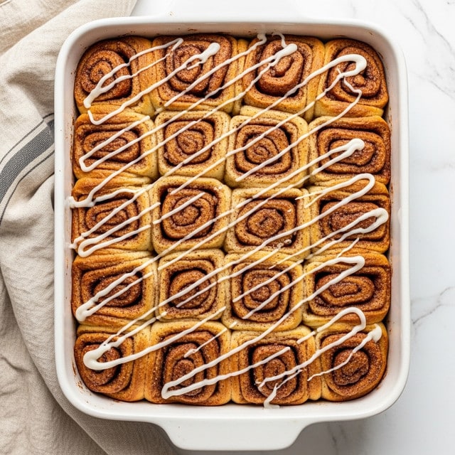 A white baking dish filled with a cinnamon pull-apart bread made of many golden-brown dough pieces coated with cinnamon sugar, layered closely together to form a textured surface. The bread is drizzled with a thin layer of white icing, creating a contrast against the warm tones of the bread. The dish rests on a white marbled surface with a beige cloth with thin dark stripes partially in view on one side. The overall look is warm, inviting, and freshly baked. photo taken with an iphone --ar 4:5 --v 7