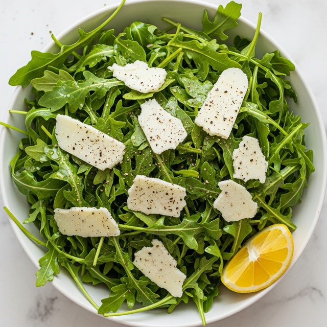 A white bowl on a white marbled surface holds a fresh arugula salad, featuring bright green leaves with a slightly glossy texture indicating light dressing. Scattered on top are thin, irregular pale yellow shavings of cheese, and some coarse black pepper is sprinkled over everything. Around the bowl, some cheese shavings and a lemon half with visible juicy pulp are set on the white marbled surface, adding context to the fresh and simple salad. Photo taken with an iphone --ar 4:5 --v 7
