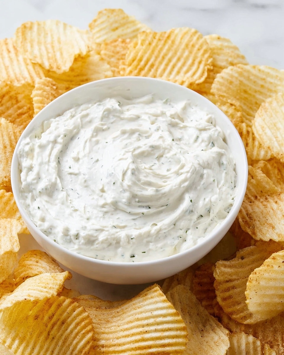 A white bowl filled with creamy, thick white dip with small green herb specks is placed at the center. Surrounding the bowl, there are many ridged potato chips in light golden yellow color, showing their crunchy texture. The scene is set on a white marbled surface, giving a clean and bright look. The chips gently lean against the bowl, creating a cozy arrangement. photo taken with an iphone --ar 4:5 --v 7