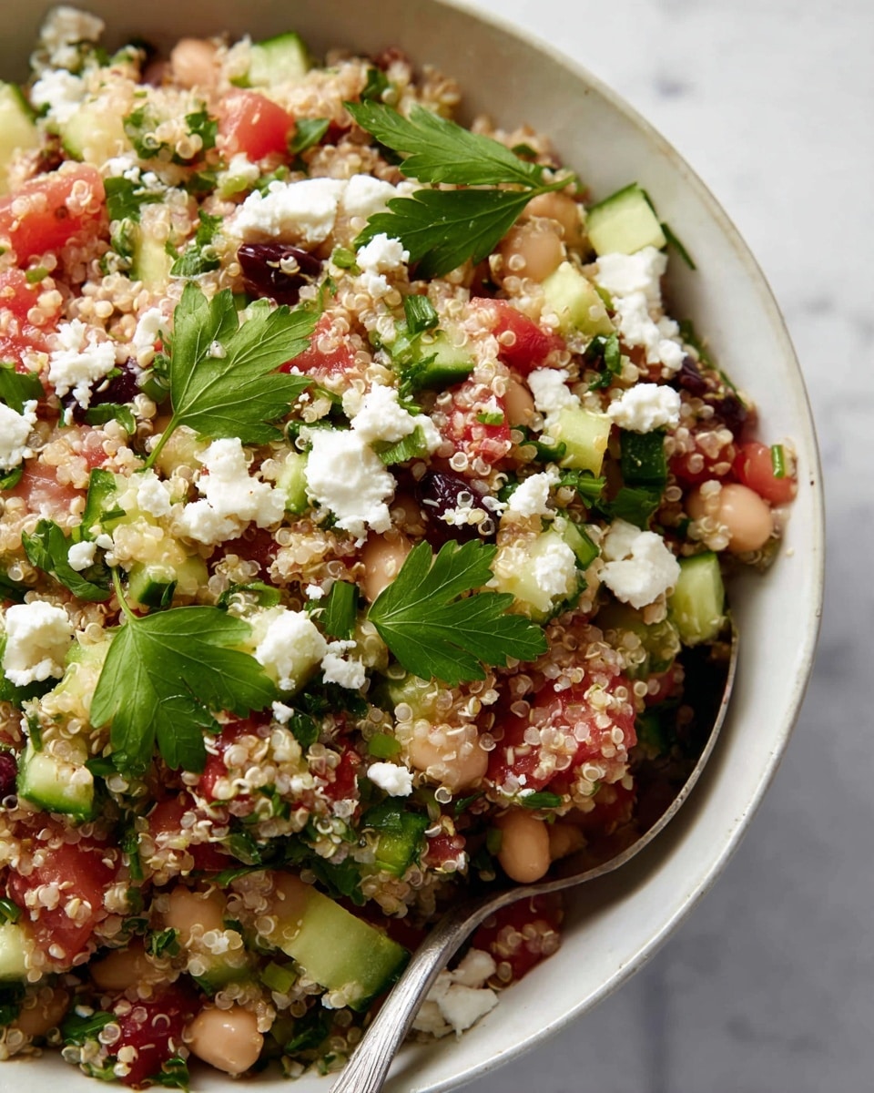 A close-up image of a quinoa salad served on an oval white plate with small black specks. The salad has multiple layers and colors: the base layer is light beige quinoa mixed with white beans, visible in soft, round shapes; scattered throughout are small red tomato pieces and dark purple-black olives. On top of this, there are small chopped green cucumber pieces and fresh leafy herbs in bright green, finely chopped and spread evenly. White crumbled cheese is sprinkled over the salad in small clusters. The plate is placed on a white marbled textured surface, with a few small purple flower buds around the edge. photo taken with an iphone --ar 4:5 --v 7