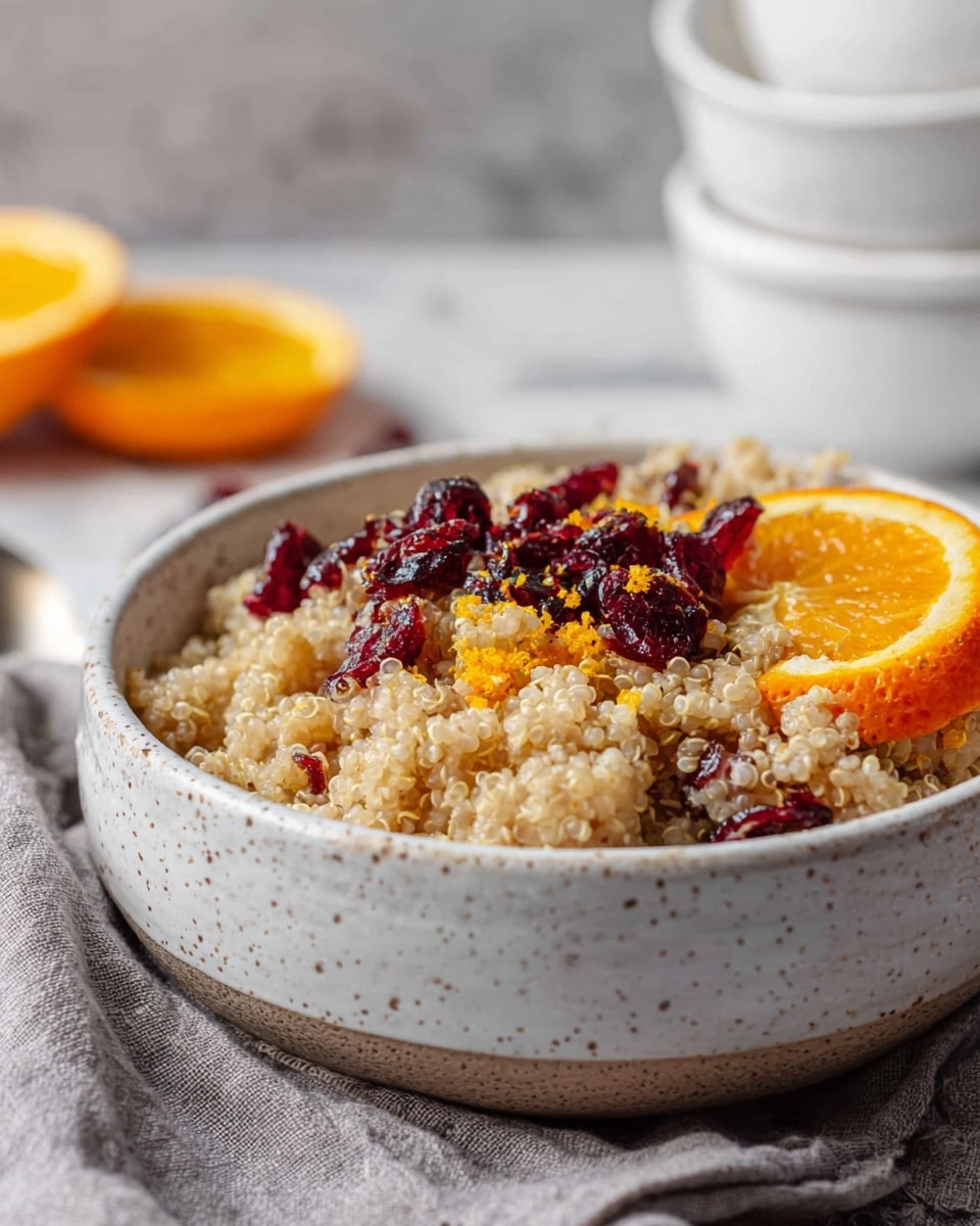 A white speckled bowl filled with creamy, light beige quinoa topped with deep red dried cranberries scattered on one side. On the other side, there is a bright orange slice and some orange zest sprinkled over the quinoa, adding small pop of color. The bowl sits on a soft grey cloth on a white marbled surface. In the blurred background, two white stacked bowls and a few orange slices can be seen. The light is soft and natural, showing the texture and colors clearly. photo taken with an iphone --ar 4:5 --v 7