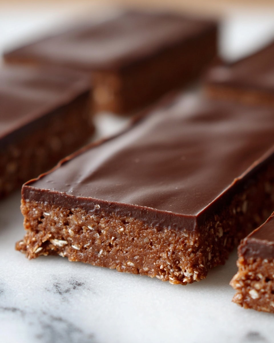 Two square pieces of chocolate bars rest on a white plate. Each bar has two visible layers: a bottom layer that is a textured, crumbly light brown base, and a smooth, dark brown chocolate top layer that is thick and glossy. One piece has a bite taken from one corner, showing the contrast between the crunchy base and the dense chocolate top. The plate is placed on a white marbled surface with a cloth featuring teal and white stripes partially visible beneath it. Photo taken with an iphone --ar 4:5 --v 7