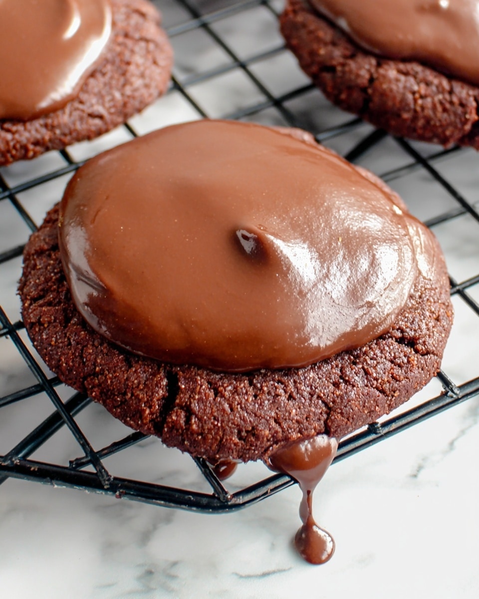 A close-up of a single chocolate cookie with one bite taken out, showing its dense, slightly crumbly texture inside. The cookie has one thick glossy layer of chocolate icing smoothly spread on top, with some parts slightly dripping over the edge. It sits on a metal spatula on a black wire cooling rack with other similar cookies blurred in the background. The surface below is white marbled texture, and a small dollop of icing drips onto the rack near the spatula. photo taken with an iphone --ar 4:5 --v 7