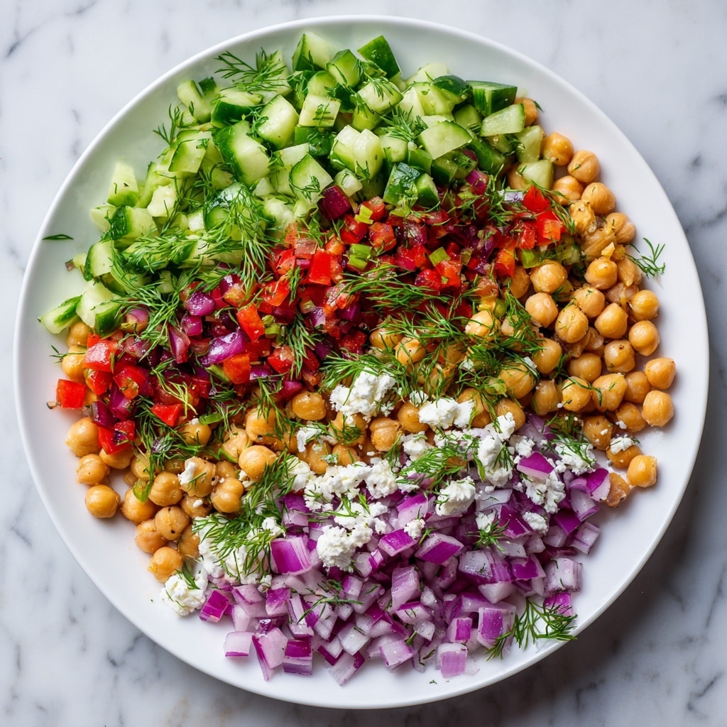 A white plate filled with a colorful salad made of three main layers: the bottom layer has small round beige chickpeas evenly spread, the middle layer consists of bright green cucumber cubes with a fresh texture, and the top layer includes small pieces of red bell pepper and finely chopped purple onion, all sprinkled with small white cheese chunks and garnished with fresh green dill leaves. The dish sits on a white marbled surface under natural light. photo taken with an iphone --ar 4:5 --v 7