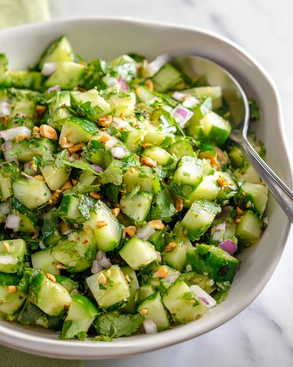 A white bowl with a wavy edge holds a fresh salad made of chopped green cucumbers, finely diced purple onions, and chopped green herbs, topped with small bits of crushed nuts. The bowl sits on a round white plate, which is surrounded by several slices of toasted bread with a golden-brown crust and soft white inside. In the background are two blue water glasses and a white marbled surface. A metal spoon rests inside the bowl. photo taken with an iphone --ar 4:5 --v 7