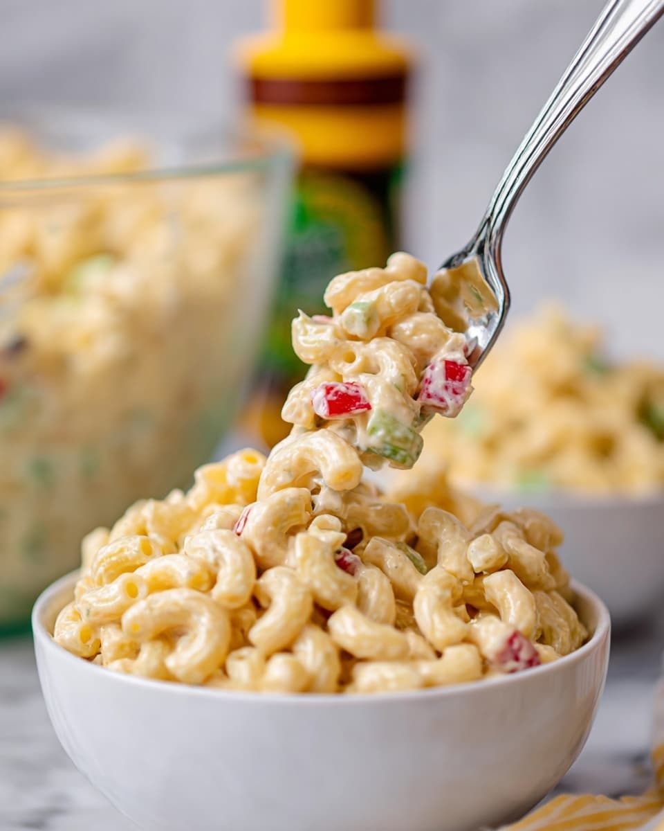 A close-up view of a white macaroni salad served in a clear glass bowl, filled with creamy, pale yellow elbow macaroni pasta mixed evenly with small chopped pieces of red bell pepper, green celery slices, and green onions. The pasta is coated with a thick, glossy white dressing that gives a rich and smooth texture. The bowl is placed on a white marbled surface with a red and white striped cloth underneath. Photo taken with an iphone --ar 4:5 --v 7