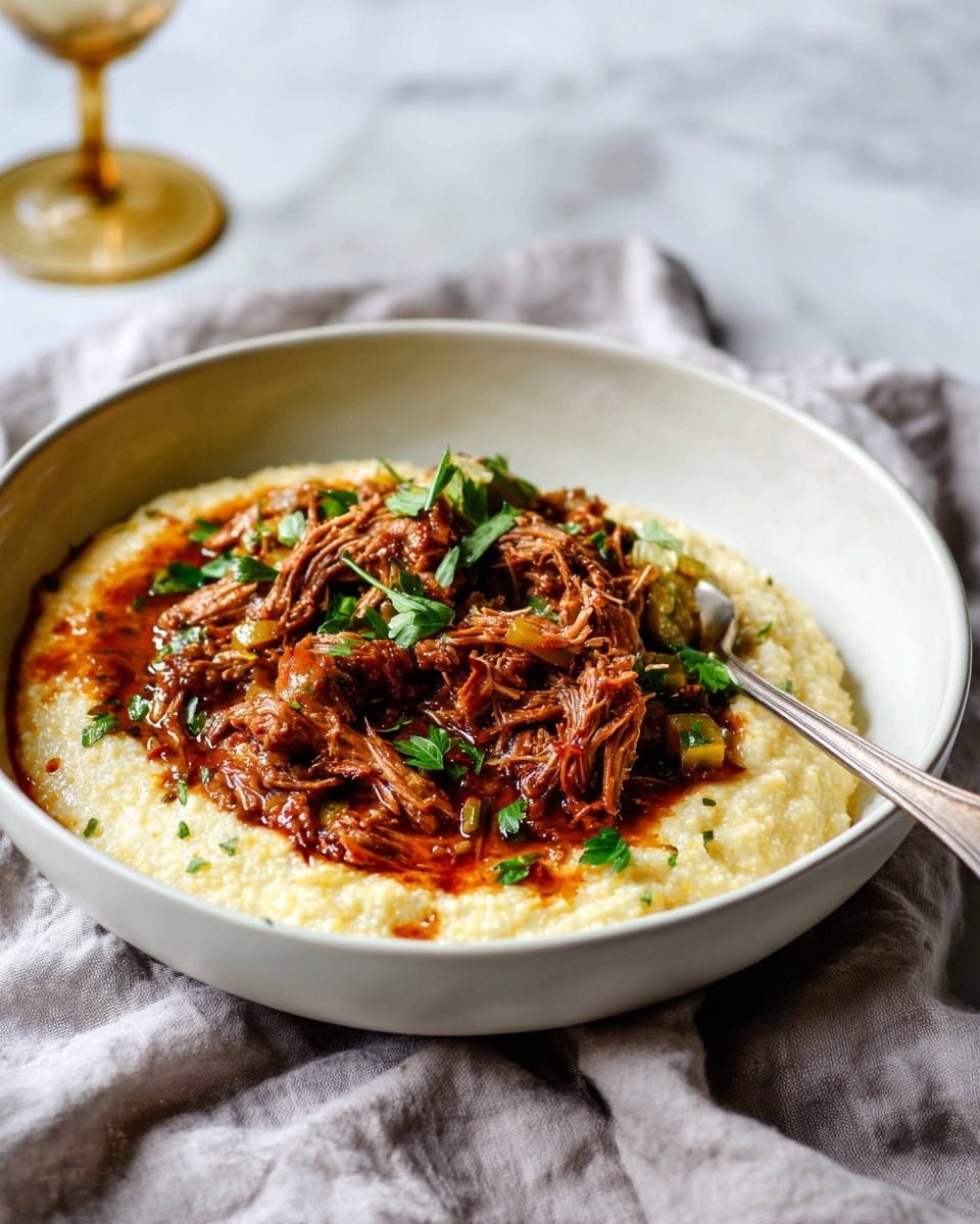 A deep white bowl holds a dish with two main layers: the bottom layer is a creamy, pale yellow porridge with a smooth, slightly grainy texture that fills the base of the bowl; on top, there is a generous pile of shredded brown meat in a thick reddish-brown sauce with visible herbs and small vegetable chunks scattered throughout, garnished with fresh green parsley leaves. A silver fork rests inside the bowl on the right side, and the bowl sits on a crumpled light gray cloth over a white marbled surface. In the blurry background, there is a glass with a gold-colored stem. photo taken with an iphone --ar 4:5 --v 7