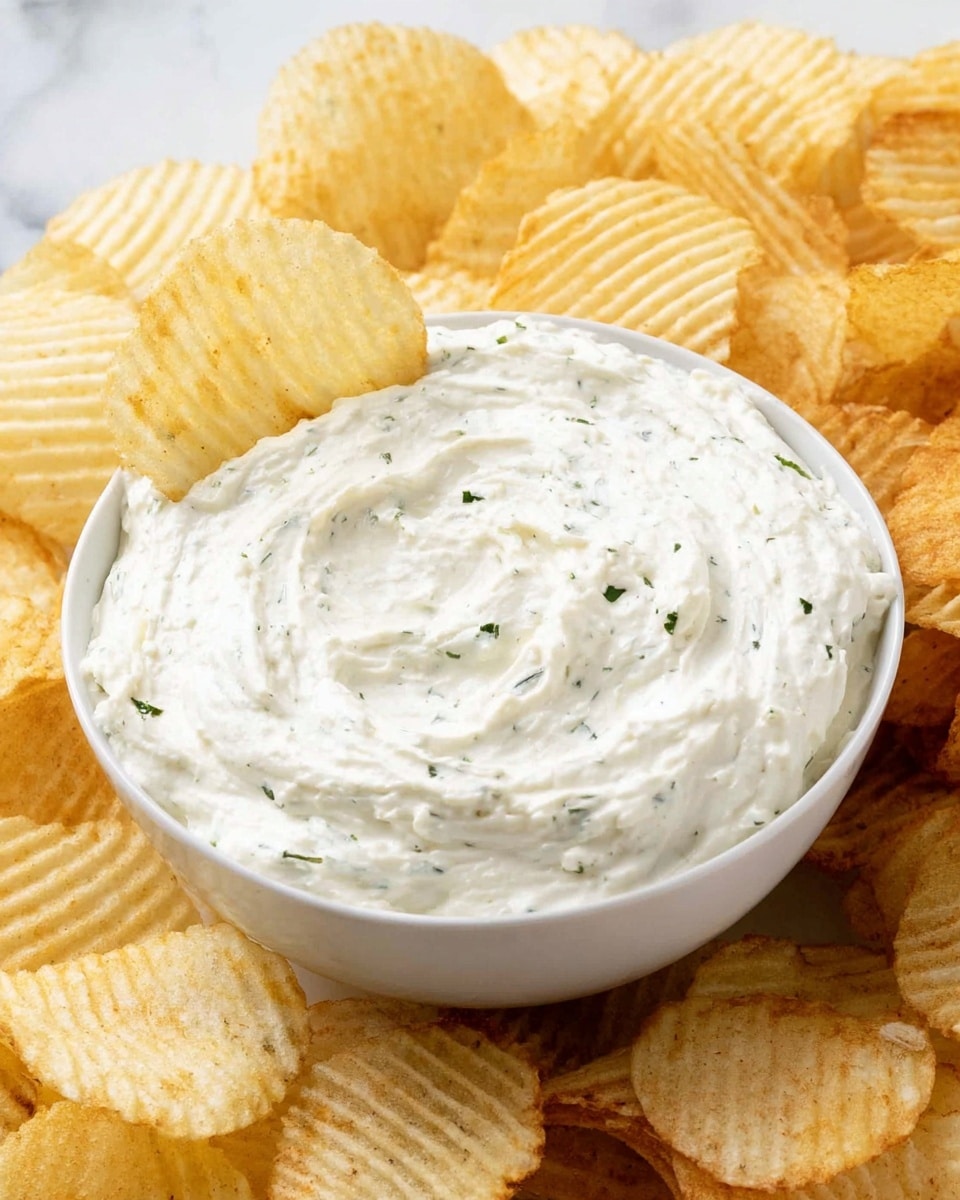 A close-up shows a woman's hand holding a single ridged potato chip dipped halfway into a white bowl filled with creamy, light-colored dip containing visible small herb bits. Surrounding the bowl are many golden ridged potato chips spread out on a white marbled surface. The dip has a smooth, thick texture with gentle swirl marks on the surface. photo taken with an iphone --ar 4:5 --v 7