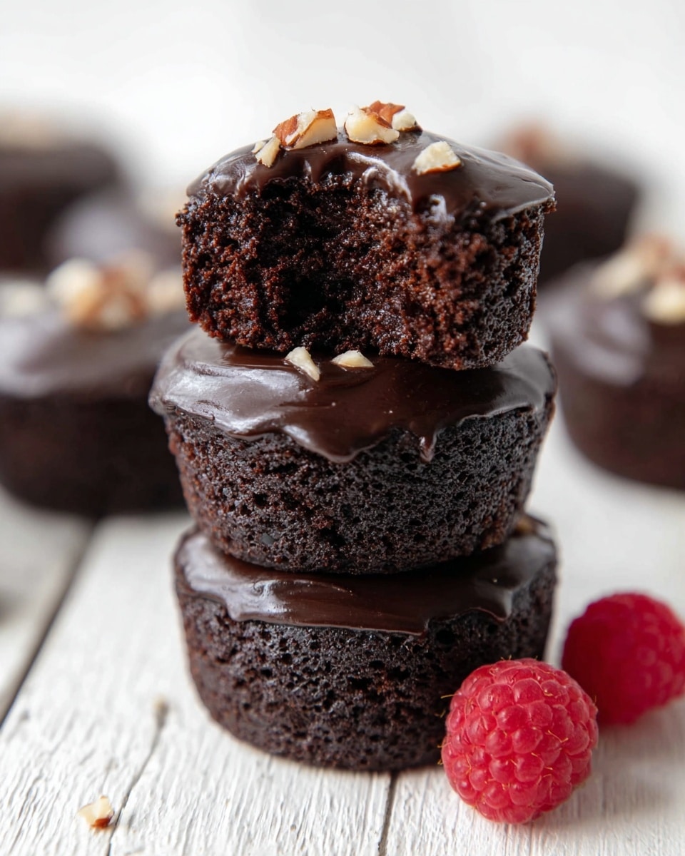 The image shows several small chocolate cakes, each with a shiny dark chocolate glaze on top, sprinkled with chopped nuts including pecans. One cake in the front is placed on a white star-shaped plate with a black pattern, sitting on a white marbled surface along with a fresh red raspberry nearby. In the background, a white rectangular plate holds more of these chocolate cakes, with some nuts on top as well, and a white bowl filled with fresh raspberries is visible. A dusty rose-colored cloth is placed under the rectangular plate, adding a soft contrast to the white marbled background. Photo taken with an iphone --ar 4:5 --v 7