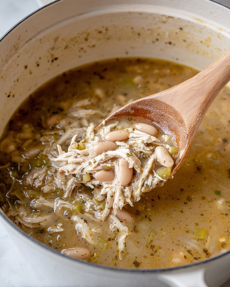 A close-up view of a white pot filled with a light brown broth soup that contains shredded chicken, white beans, and small pieces of green vegetables. The soup has a slightly oily surface with specks of black pepper and herbs. A wooden spoon is lifting a spoonful of the soup, showing tender shredded chicken mixed with beans and broth. The inside walls of the pot have some splashes of soup. The pot sits on a white marbled textured surface. photo taken with an iphone --ar 4:5 --v 7
