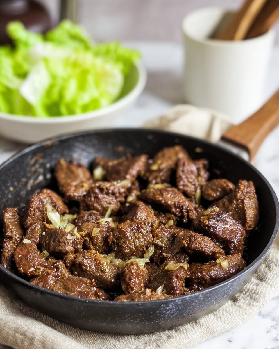 The image shows a black pan filled with several pieces of cooked meat that are browned and textured, with small bits of cooked onions scattered throughout. The meat pieces are uneven in size and shape, with a slightly crispy surface and some glistening from cooking juices. In the background, there is a blurred bunch of green lettuce leaves adding a fresh green color. At the front, there is a small white bowl filled with coarse salt and a wooden spoon resting on top. The scene is set on a white marbled texture surface with a light-colored cloth partly visible under the pan. photo taken with an iphone --ar 4:5 --v 7