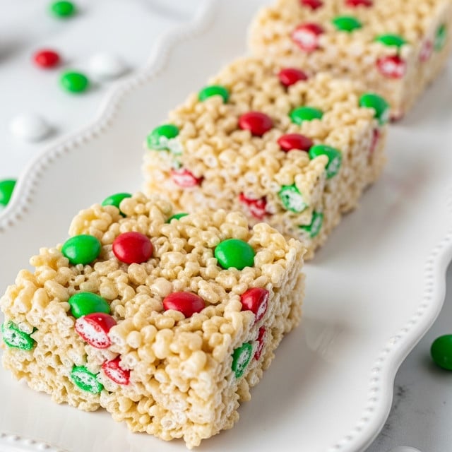 Three square crispy rice treats sit on a white plate with a scalloped edge, arranged in a diagonal line. Each treat is light beige with a glossy, sticky texture, filled with small red and green candy bits scattered evenly throughout. The treats look thick and soft, with a slightly uneven surface due to the puffed rice and candy mix. The background shows a smooth white marbled texture and some small round candies in a pale green color nearby. Photo taken with an iphone --ar 4:5 --v 7