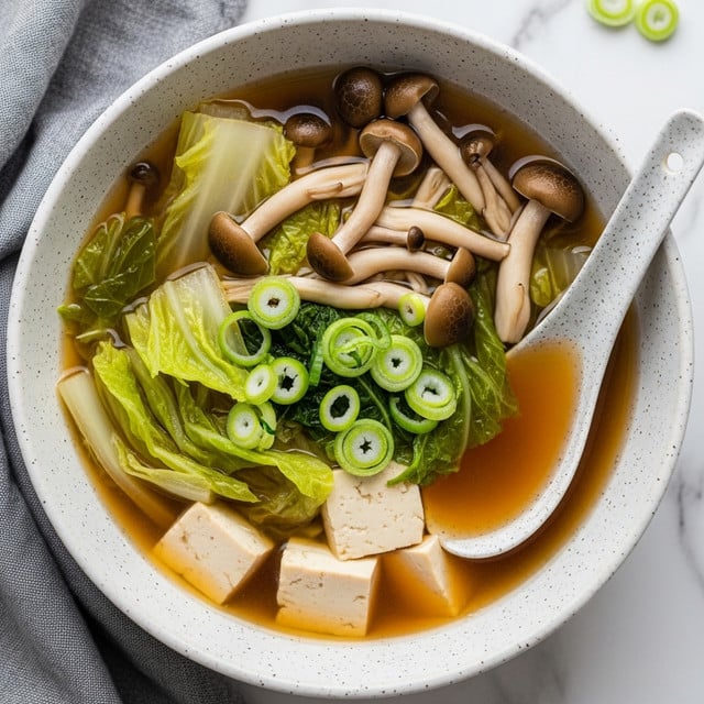 The image shows a white speckled bowl filled with a light brown broth that holds several layers of ingredients. On the bottom layer, there are pale beige tofu cubes and light green pieces of cabbage submerged in the broth. On top, brown shimeji mushrooms with smooth caps and stalks float among the cabbage leaves. Thin, round green scallion slices are scattered over the surface, adding a pop of bright green color. The bowl is accompanied by a white speckled spoon resting inside and is set on a white marbled table with a gray cloth nearby. Photo taken with an iphone --ar 4:5 --v 7