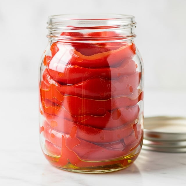 A clear glass jar with a white lid is filled with several layers of soft, peeled roasted red pepper slices. The peppers are bright red with a slightly shiny, moist texture, tightly packed inside the jar. At the bottom, there is a small layer of golden olive oil that adds a glossy look. The jar sits on a white marbled surface, and the background is softly lit with white light. photo taken with an iphone --ar 4:5 --v 7