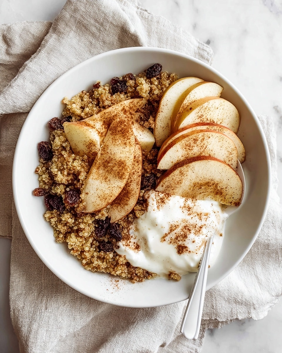 A white bowl with a base layer of cooked quinoa mixed with dark raisins, giving a grainy light brown and dark contrast. On one side of the bowl, there is a curved row of thinly sliced pale apple pieces dusted with brown cinnamon powder. Overlapping the apple slices, there is a portion of creamy white yogurt also sprinkled with cinnamon. A white spoon is placed inside the bowl, slightly scooping the yogurt and quinoa mix. The bowl sits on a beige linen cloth over a white marbled texture. Photo taken with an iphone --ar 4:5 --v 7