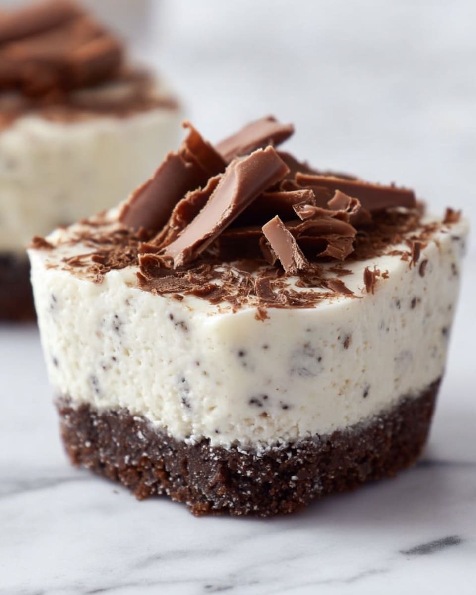 The image shows small dessert cups with two distinct layers on a white marbled surface. The bottom layer is dark brown, dense, and crumbly, resembling a chocolate cookie crust. The top layer is thick, creamy, and white with small dark chocolate flakes mixed throughout, giving it a textured look. On the very top, there are curled dark brown chocolate shavings scattered lightly, adding a decorative touch. The focus is on the front dessert cup, with two more similar cups blurred in the background. photo taken with an iphone --ar 4:5 --v 7